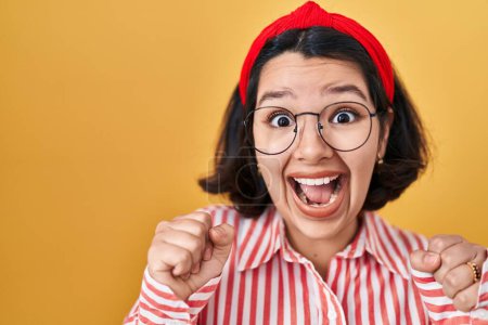 Téléchargez les photos : Jeune femme hispanique portant des lunettes sur fond jaune criant fier, célébrant la victoire et le succès très excité avec les bras levés - en image libre de droit