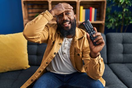 Foto de Hombre afroamericano sosteniendo control remoto de televisión estresado y frustrado con la mano en la cabeza, cara sorprendida y enojada - Imagen libre de derechos