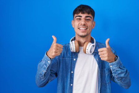 Foto de Young hispanic man standing over blue background success sign doing positive gesture with hand, thumbs up smiling and happy. cheerful expression and winner gesture. - Imagen libre de derechos