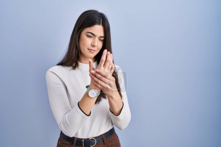 Téléchargez les photos : Jeune femme brune debout sur fond bleu souffrant de douleur aux mains et aux doigts, inflammation de l'arthrite - en image libre de droit