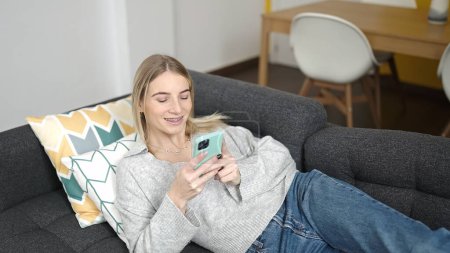 Téléchargez les photos : Jeune femme blonde utilisant un smartphone couché sur le canapé à la maison - en image libre de droit