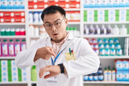 Téléchargez les photos : Jeune homme chinois travaillant à la pharmacie pharmacie à la hâte pointant vers regarder le temps, l'impatience, bouleversé et en colère pour retard de délai - en image libre de droit