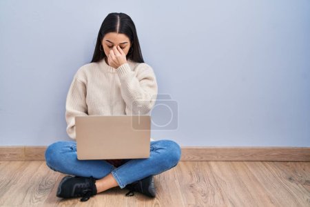 Young woman using laptop sitting on the floor at home tired rubbing nose and eyes feeling fatigue and headache. stress and frustration concept. 