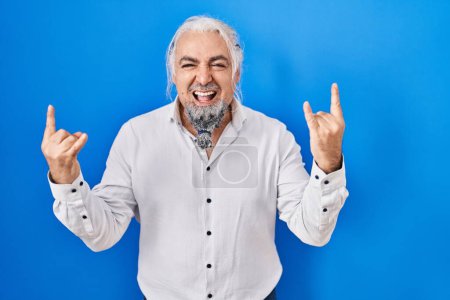 Photo for Middle age man with grey hair standing over blue background shouting with crazy expression doing rock symbol with hands up. music star. heavy music concept. - Royalty Free Image