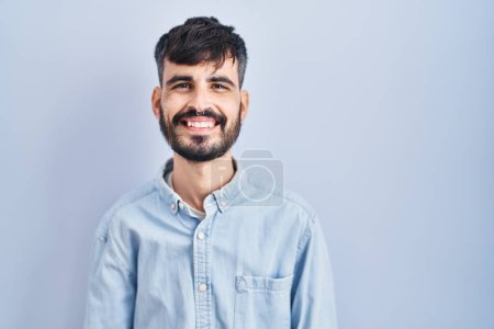Foto de Joven hombre hispano con barba de pie sobre fondo azul con una sonrisa alegre y fresca en la cara. persona afortunada. - Imagen libre de derechos