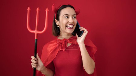 Téléchargez les photos : Young beautiful hispanic woman wearing devil costume talking on smartphone over isolated red background - en image libre de droit