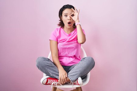 Foto de Mujer joven hispana sentada en una silla sobre fondo rosa haciendo un gesto bien sorprendido con la cara sorprendida, los ojos mirando a través de los dedos. expresión incrédula. - Imagen libre de derechos