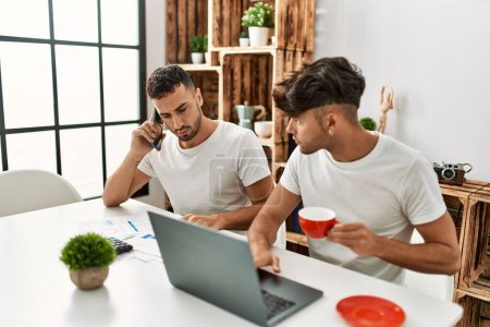 Foto de Dos hombres hispanos en pareja hablando en el teléfono inteligente y usando un portátil trabajando en casa - Imagen libre de derechos