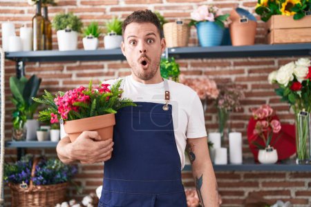 Foto de Joven hombre hispano trabajando en floristería sosteniendo maceta asustado y sorprendido con la boca abierta para sorpresa, cara de incredulidad - Imagen libre de derechos