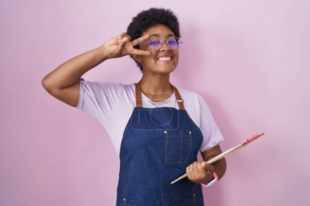Téléchargez les photos : Jeune femme afro-américaine portant tablier de serveuse professionnel tenant presse-papiers faisant symbole de paix avec les doigts sur le visage, souriant joyeuse montrant la victoire - en image libre de droit