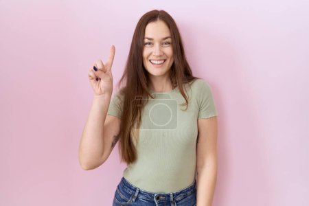 Téléchargez les photos : Belle femme brune debout sur fond rose montrant et pointant vers le haut avec le doigt numéro un tout en souriant confiant et heureux. - en image libre de droit