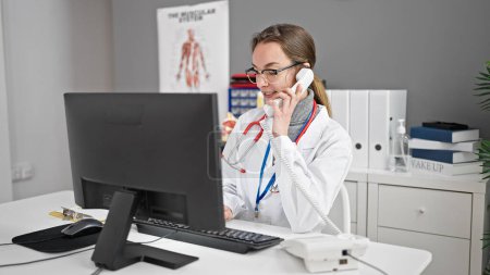 Téléchargez les photos : Jeune femme caucasienne médecin utilisant l'ordinateur parlant au téléphone à la clinique - en image libre de droit