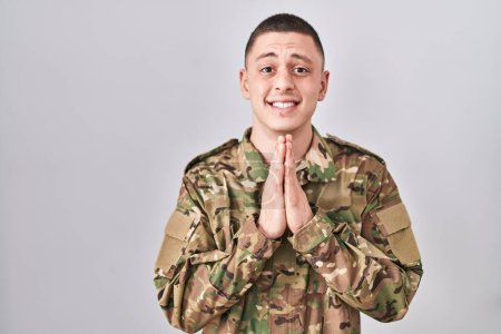 Foto de Joven vestido con camuflaje uniforme del ejército rezando con las manos juntas pidiendo perdón sonriendo confiado. - Imagen libre de derechos