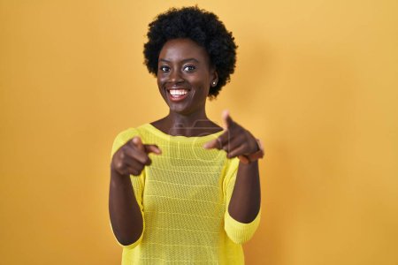 Photo for African young woman standing over yellow studio pointing to you and the camera with fingers, smiling positive and cheerful - Royalty Free Image