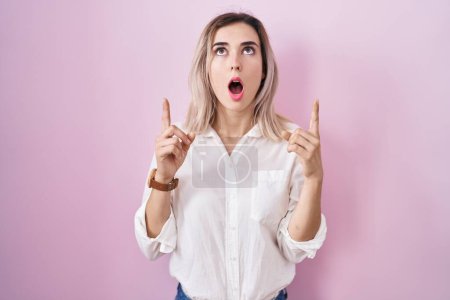 Foto de Young beautiful woman standing over pink background amazed and surprised looking up and pointing with fingers and raised arms. - Imagen libre de derechos