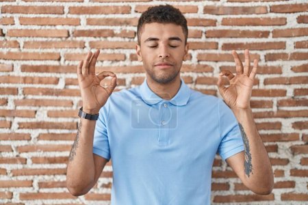 Téléchargez les photos : Jeune homme brésilien debout sur un mur de briques détendu et souriant les yeux fermés en faisant un geste de méditation avec les doigts. concept de yoga. - en image libre de droit