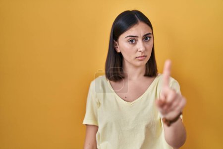 Foto de Chica hispana vistiendo camiseta casual sobre fondo amarillo apuntando con el dedo hacia arriba y expresión enojada, sin mostrar ningún gesto - Imagen libre de derechos