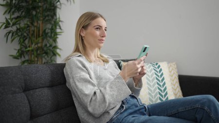Téléchargez les photos : Jeune femme blonde utilisant un smartphone assis sur le canapé à la maison - en image libre de droit