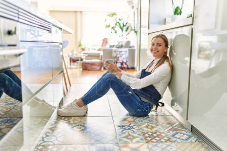 Téléchargez les photos : Jeune femme blonde buvant du café assis sur le sol à la cuisine - en image libre de droit