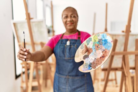 Foto de Mujer afroamericana mayor sonriendo confiada sosteniendo paleta y pincel en el estudio de arte - Imagen libre de derechos