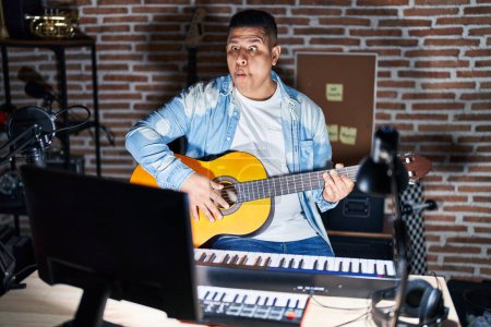 Photo for Hispanic young man playing classic guitar at music studio making fish face with lips, crazy and comical gesture. funny expression. - Royalty Free Image