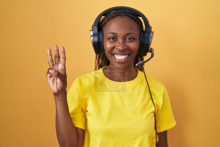 Foto de Mujer afroamericana escuchando música usando auriculares mostrando y señalando con los dedos número tres mientras sonríe confiada y feliz. - Imagen libre de derechos