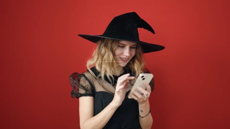 Téléchargez les photos : Jeune femme blonde portant un costume de sorcière en utilisant un smartphone sur fond rouge isolé - en image libre de droit