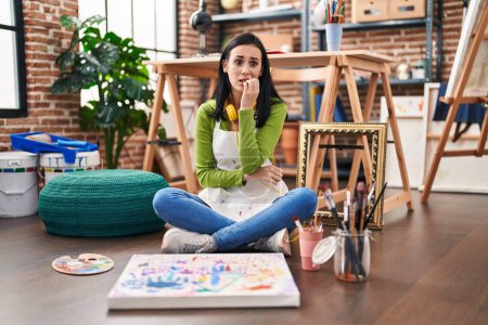 Téléchargez les photos : Femme hispanique assise au studio d'art peignant sur toile regardant stressé et nerveux avec les mains sur les ongles mordants de la bouche. problème d'anxiété. - en image libre de droit