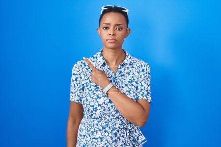 Photo for African american woman standing over blue background pointing with hand finger to the side showing advertisement, serious and calm face - Royalty Free Image