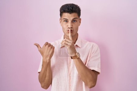 Foto de Young hispanic man standing over pink background asking to be quiet with finger on lips pointing with hand to the side. silence and secret concept. - Imagen libre de derechos