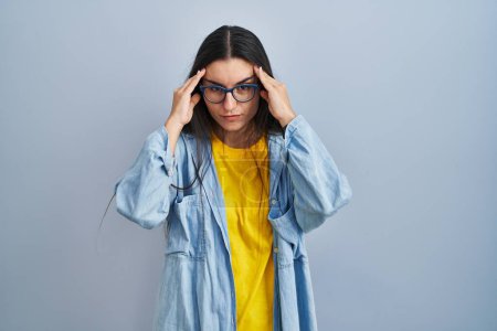 Foto de Mujer hispana joven de pie sobre fondo azul con la mano en la cabeza, dolor de cabeza porque el estrés. padecer migraña. - Imagen libre de derechos