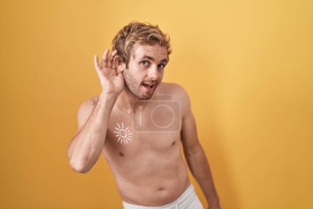 Téléchargez les photos : Homme caucasien debout torse nu portant un écran solaire souriant avec la main sur l'oreille en écoutant une rumeur ou des commérages. concept de surdité. - en image libre de droit