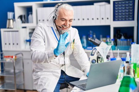 Téléchargez les photos : Homme d'âge moyen avec les cheveux gris travaillant au laboratoire scientifique faire appel vidéo regardant positif et heureux debout et souriant avec un sourire confiant montrant les dents - en image libre de droit