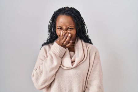 Foto de Mujer africana de pie sobre fondo blanco oliendo algo apestoso y asqueroso, olor intolerable, conteniendo la respiración con los dedos en la nariz. mal olor - Imagen libre de derechos
