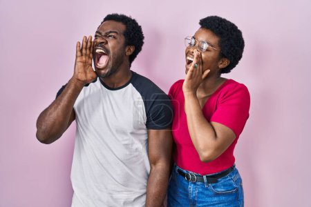 Foto de Joven pareja afroamericana de pie sobre fondo rosa gritando y gritando fuerte a un lado con la mano en la boca. concepto de comunicación. - Imagen libre de derechos