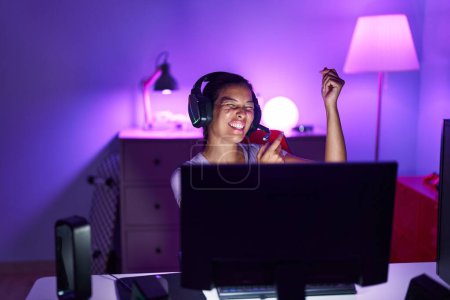 Foto de Joven hermosa mujer hispana streamer jugando videojuego con la expresión ganadora en la sala de juegos - Imagen libre de derechos