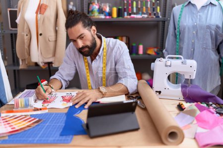 Foto de Joven hombre hispano sastrería dibujo ropa diseño usando touchpad en atelier - Imagen libre de derechos