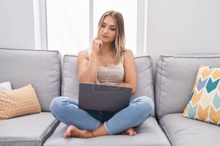 Téléchargez les photos : Blonde femme caucasienne utilisant un ordinateur portable à la maison assis sur le canapé visage sérieux penser à la question avec la main sur le menton, réfléchi sur l'idée déroutante - en image libre de droit