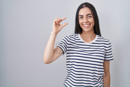 Téléchargez les photos : Jeune femme brune portant t-shirt rayé geste souriant et confiant avec la main faisant signe de petite taille avec les doigts à la recherche et la caméra. concept de mesure. - en image libre de droit