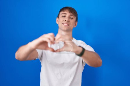 Photo for Caucasian blond man standing over blue background smiling in love doing heart symbol shape with hands. romantic concept. - Royalty Free Image