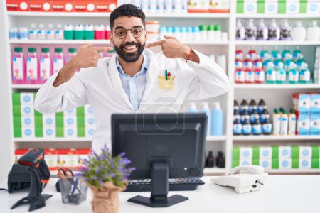Téléchargez les photos : Homme hispanique avec barbe travaillant à la pharmacie pharmacie souriant gai montrant et pointant du doigt les dents et la bouche. concept de santé dentaire. - en image libre de droit