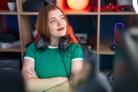 Téléchargez les photos : Jeune rousse femme streamer souriant confiant assis avec les bras croisés geste à la salle de jeux - en image libre de droit