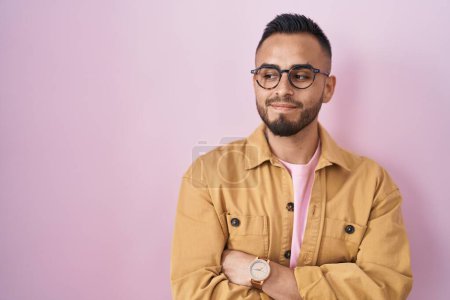 Foto de Joven hombre hispano de pie sobre fondo rosa sonriendo mirando hacia un lado y mirando hacia otro pensando. - Imagen libre de derechos
