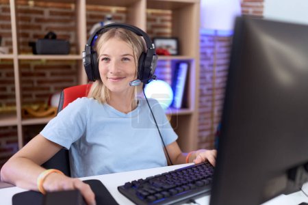 Téléchargez les photos : Jeune femme caucasienne jouant à des jeux vidéo portant des écouteurs regardant de côté avec le sourire sur le visage, expression naturelle. riant confiant. - en image libre de droit