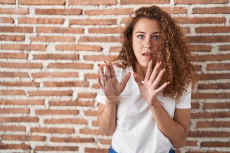 Photo for Young caucasian woman standing over bricks wall background afraid and terrified with fear expression stop gesture with hands, shouting in shock. panic concept. - Royalty Free Image