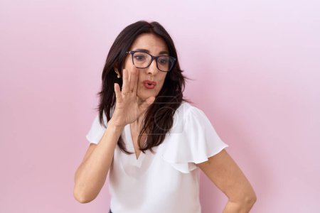 Téléchargez les photos : Moyen Age femme hispanique portant casual t-shirt blanc et lunettes main sur la bouche racontant rumeur secrète, chuchotant conversation malicieuse - en image libre de droit