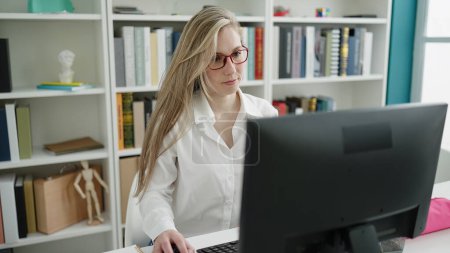 Téléchargez les photos : Jeune étudiante blonde utilisant un ordinateur assis sur la table à la bibliothèque de l'université - en image libre de droit