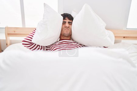 Photo for Young hispanic man covering ears with pillow for noise at bedroom - Royalty Free Image
