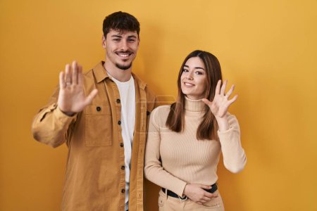 Téléchargez les photos : Jeune couple hispanique debout sur fond jaune renonçant à dire bonjour heureux et souriant, geste de bienvenue amical - en image libre de droit
