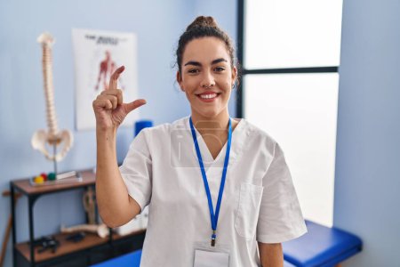 Téléchargez les photos : Jeune femme hispanique travaillant à la clinique de réadaptation geste souriant et confiant avec la main faisant signe de petite taille avec les doigts regardant et la caméra. concept de mesure. - en image libre de droit